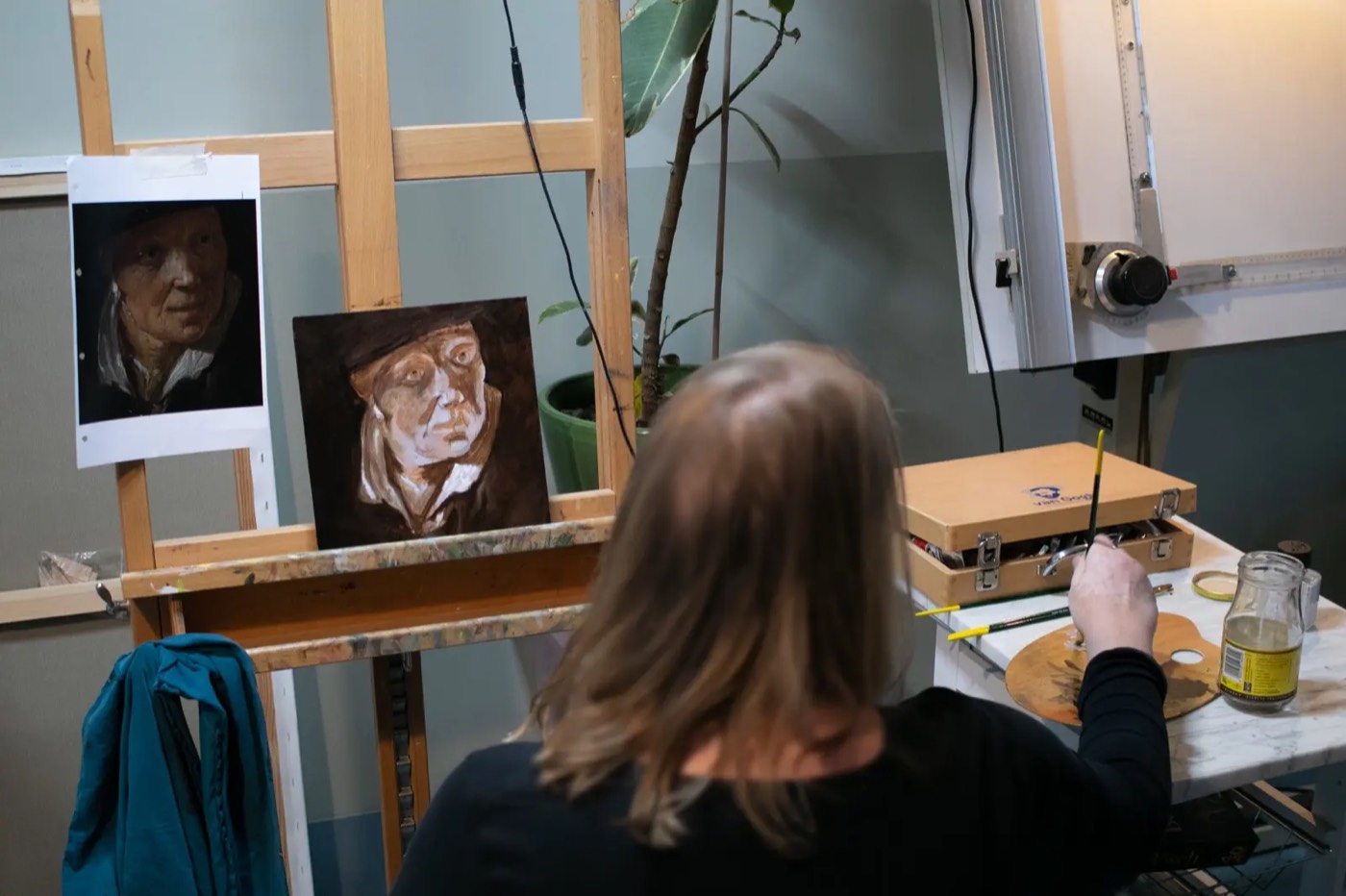 A student at the easel painting a portrait in oil, the photograph she is working from pinned up beside it.